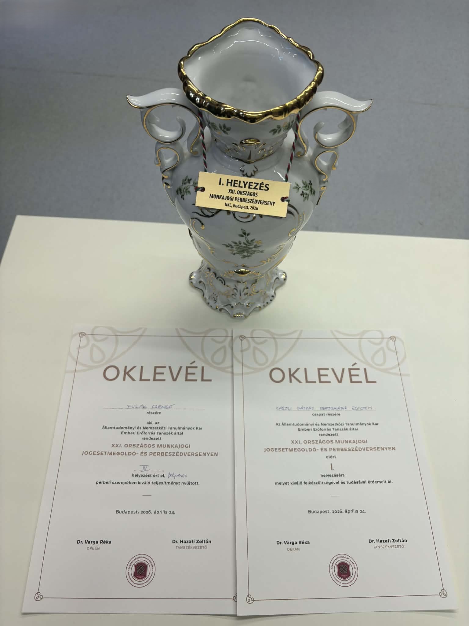 A képen a 2 db oklevél látható és az első helyezésért járó hollóházi barokk Erika mintás váza, amelyet a XXI. Országos Munkajogi Jogesetmegoldó- és Perbeszédversenyen kaptak a KRE ÁJK hallgatói.